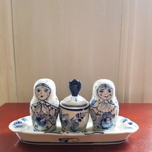 Könnte beinhalten: Ein weißes Salz- und Pfefferstreuer-Set aus Keramik mit einem passenden Tablett. Die Streuer sind mit blauen und weißen Blumenmustern verziert und zeigen die Gesichter von russischen Matrjoschka-Puppen.