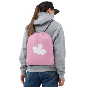 May include: A pink drawstring backpack with a white maple leaf emblem. The bag has black drawstrings and is worn over the shoulders. The bag is made of a soft material.