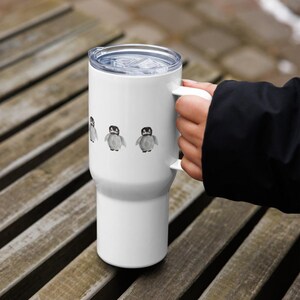 May include: White travel mug with a clear lid and handle. The mug features three watercolor penguin illustrations in black, gray, and white. The mug is designed to keep beverages hot or cold.