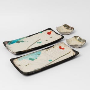 Könnte beinhalten: Ein Set aus vier Keramikgeschirren. Zwei rechteckige Teller mit schwarzen Rändern und abstrakten Designs in Blau, Grün und Rot. Zwei kleine, muschelförmige Schalen mit goldenen Akzenten.