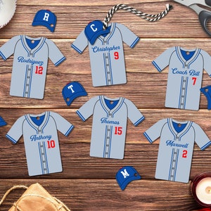 May include: Set of ten blue and white baseball jersey-shaped ornaments with red numbers and names. The ornaments include: Rodriguez 12, Christopher 9, Coach Bill 7, Anthony 10, Thomas 15, Maxwell 2.