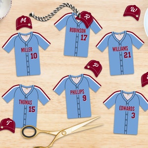 May include: Light blue baseball jersey cutouts with white and maroon accents, featuring player names and numbers. Includes matching maroon baseball caps. A baseball and a cup of coffee are also in the image.