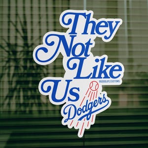 Puede incluir: Una pegatina blanca con texto azul que dice "They Not Like Us Dodgers". La pegatina tiene un gráfico de béisbol y detalles en rojo. La pegatina está sobre un fondo verde.