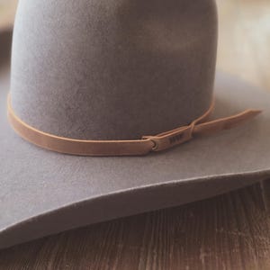 May include: A gray fedora-style hat with a wide brim and a light brown leather band. The band is secured with a small loop and has the text "1939" embossed on it. The hat is resting on a wooden surface.
