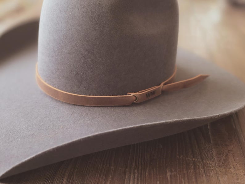 May include: A gray fedora-style hat with a wide brim and a light brown leather band. The band is secured with a small loop and has the text "1939" embossed on it. The hat is resting on a wooden surface.