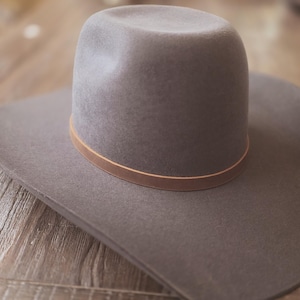 May include: A gray cowboy hat with a wide brim and a rounded crown. The hat features a thin, light brown leather band around the base of the crown. The hat is made of a soft material and is set against a wooden background.