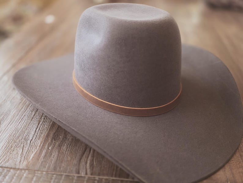 May include: A gray cowboy hat with a wide brim and a rounded crown. The hat features a thin, light brown leather band around the base of the crown. The hat is made of a soft material and is set against a wooden background.