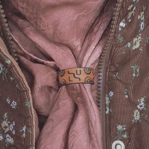 May include: A brown scarf ring with a geometric design, worn with a pink scarf and a brown floral jacket. The ring has a tan background with black and turquoise accents. The jacket is quilted and has a floral pattern.