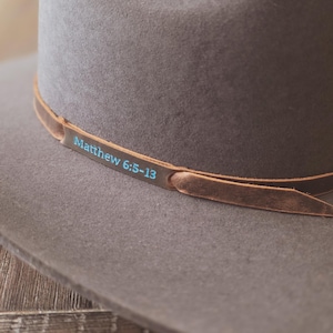 May include: A gray cowboy hat with a brown leather band. The band features the text "Matthew 6:5-13" in a teal color. The hat is made of a soft material and has a classic design.