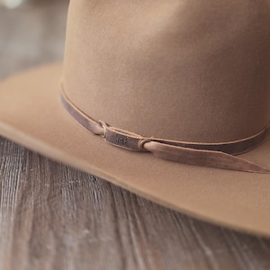 May include: A tan-colored cowboy hat with a brown leather band. The hat has a wide brim and a rounded crown. The leather band is secured with a knot and features a small embossed design.