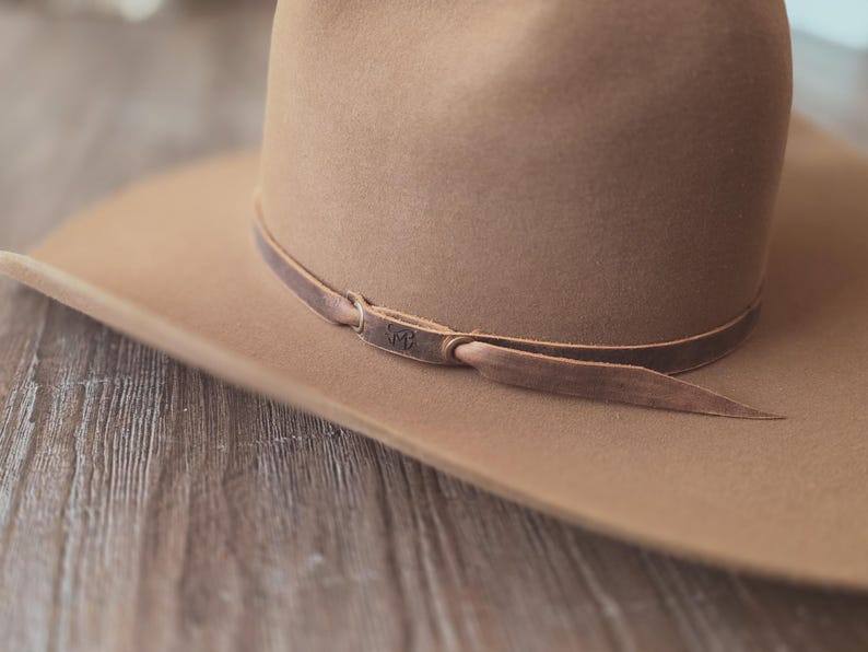 May include: A tan-colored cowboy hat with a brown leather band. The hat has a wide brim and a rounded crown. The leather band is secured with a knot and features a small embossed design.