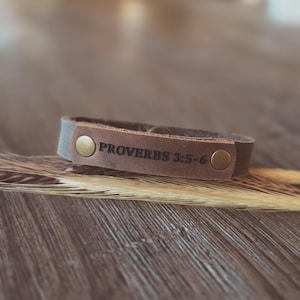 May include: A brown leather bracelet, inscribed with "PROVERBS 3:5-6", is fastened with two gold-coloured press studs. The bracelet rests on a wooden surface with dried wheat stalks.