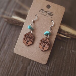 May include: Earrings with a geometric design and turquoise accents. The earrings are made of light brown leather and feature a silver hook. The earrings are displayed on a brown card with the text "Owl West Leathercraft".