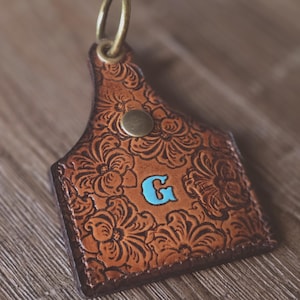 May include: Brown leather keychain with a floral embossed design. The keychain features a turquoise letter "G" and a brass-colored metal ring and rivet. The leather has a distressed look.