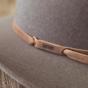 May include: Close-up of a gray fedora hat with a light brown leather band. The leather band is secured with metal eyelets and features the initials "WW" embossed on it. The hat has a wide brim and a classic design.