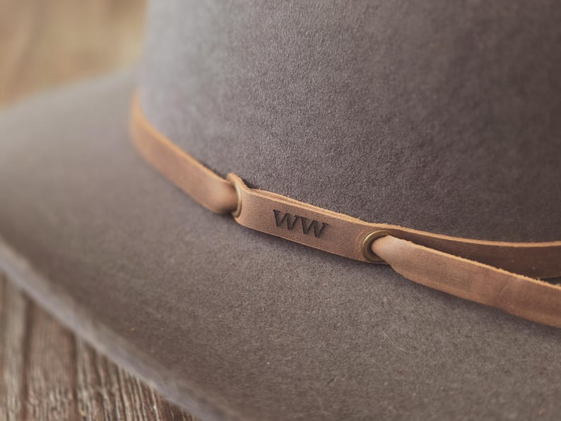 May include: Close-up of a gray fedora hat with a light brown leather band. The leather band is secured with metal eyelets and features the initials "WW" embossed on it. The hat has a wide brim and a classic design.