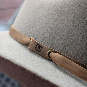 May include: A light gray fedora hat with a brown leather band. The band features the initials "DS" and is secured with metal eyelets. The hat has a classic, structured shape, suitable for various occasions.