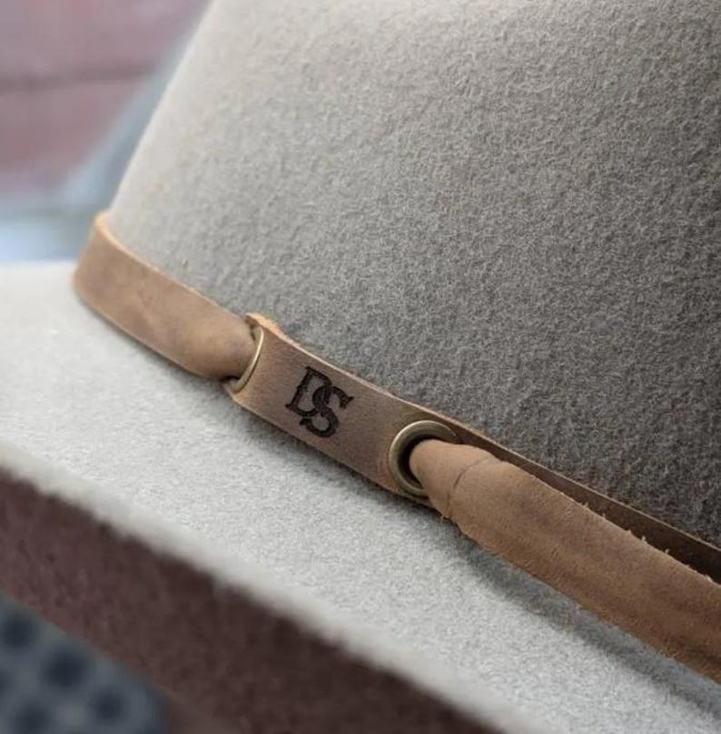 May include: A light gray fedora hat with a brown leather band. The band features the initials "DS" and is secured with metal eyelets. The hat has a classic, structured shape, suitable for various occasions.