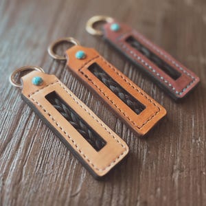 May include: Three leather keyrings in tan, orange, and burgundy, each with a braided insert and a turquoise accent. The keyrings have a rectangular shape with a stitched border and a metal ring for keys.