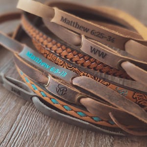 May include: A collection of leather guitar straps in various shades of brown. Some straps feature embossed designs, turquoise accents, and text including "Matthew 6:25-34" and "Matthew 6:5-13". The straps are arranged on a wooden surface.