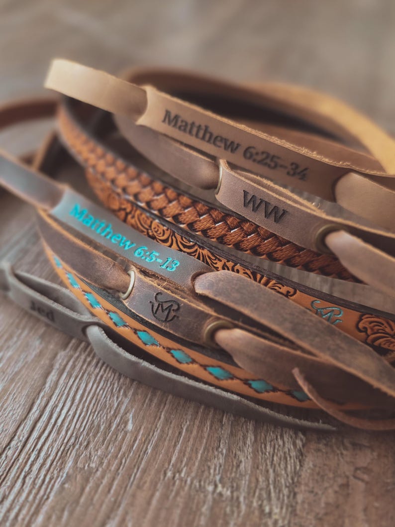 May include: A collection of leather guitar straps in various shades of brown. Some straps feature embossed designs, turquoise accents, and text including "Matthew 6:25-34" and "Matthew 6:5-13". The straps are arranged on a wooden surface.