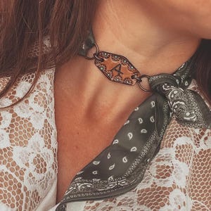 May include: A leather choker necklace with the letter "A" in the center. The choker is connected to a dark green paisley bandana. The person is wearing a white lace top.