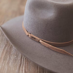 May include: A gray cowboy hat with a wide brim and a leather band. The leather band is light brown and has the letters "WW" stamped on it. The hat is made of a soft material and sits on a wooden surface.