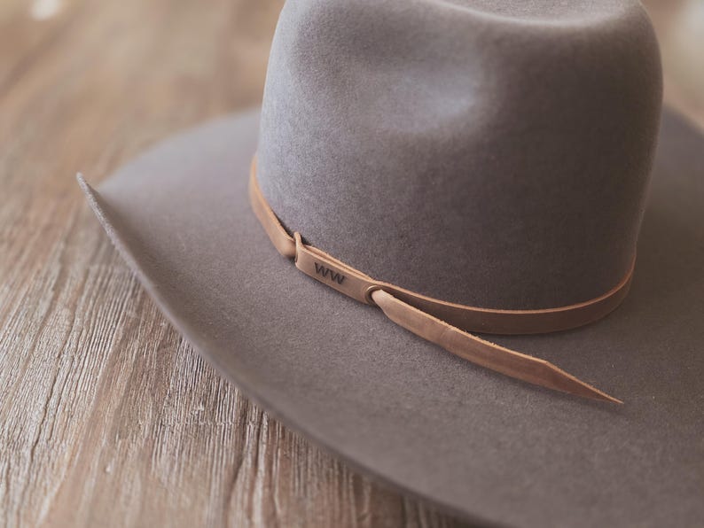 May include: A gray cowboy hat with a wide brim and a leather band. The leather band is light brown and has the letters "WW" stamped on it. The hat is made of a soft material and sits on a wooden surface.