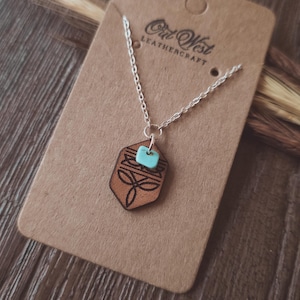 May include: A silver chain necklace with a brown leather pendant. The pendant has a geometric design and a small turquoise accent. The necklace is displayed on a brown card with the text "Out West Leathercraft".