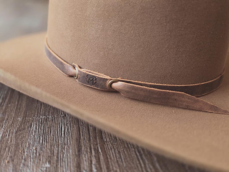 May include: A tan-colored fedora hat with a brown leather band. The hat has a wide brim and a classic shape. The leather band is secured with a knot and features a small embossed design. The hat is made of a soft material.
