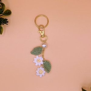 May include: A white crocheted flower keychain with green leaves and a gold key ring. The keychain has a pearl bead accent.