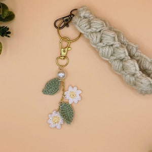 May include: A beige crocheted keychain with a gold key ring and a charm featuring white crocheted flowers and green leaves with a pearl bead.