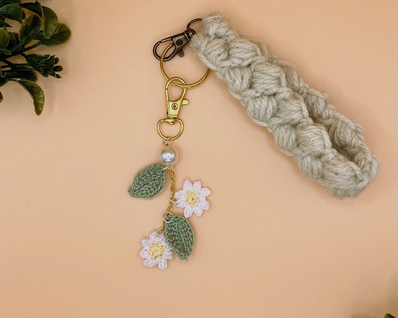 May include: A beige crocheted keychain with a gold key ring and a charm featuring white crocheted flowers and green leaves with a pearl bead.