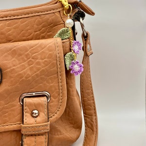 May include: A brown leather handbag with a gold chain and purple crocheted flower charm. The charm has a pearl bead and green leaves.