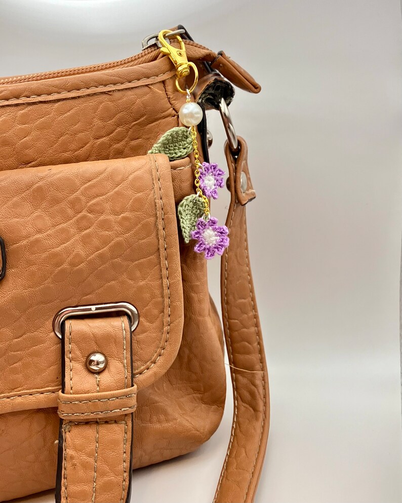 May include: A brown leather handbag with a gold chain and purple crocheted flower charm. The charm has a pearl bead and green leaves.