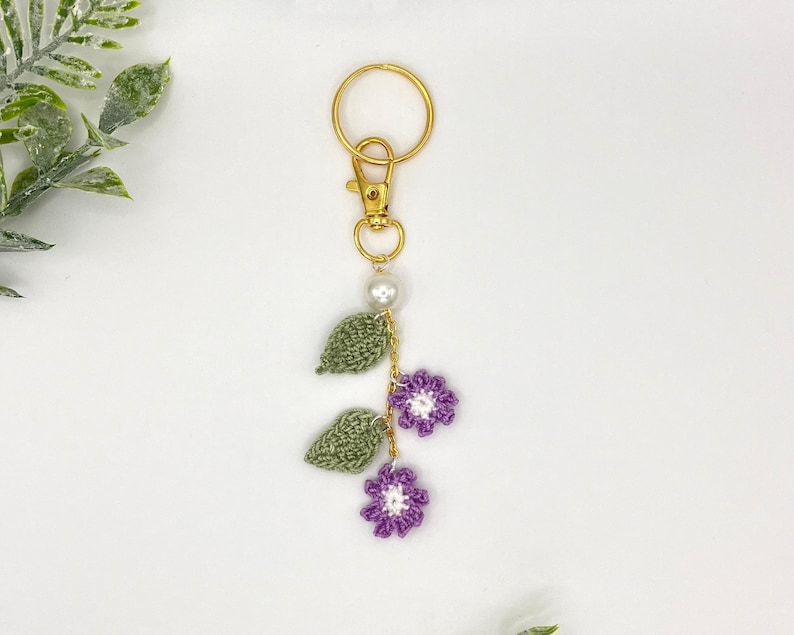 May include: A crocheted flower and leaf keychain with a gold key ring and a pearl bead. The flowers are purple and white, and the leaves are green.