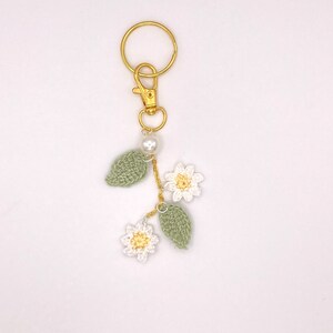 May include: A gold key chain with a crocheted flower and leaf charm. The charm is white and green with a pearl bead.
