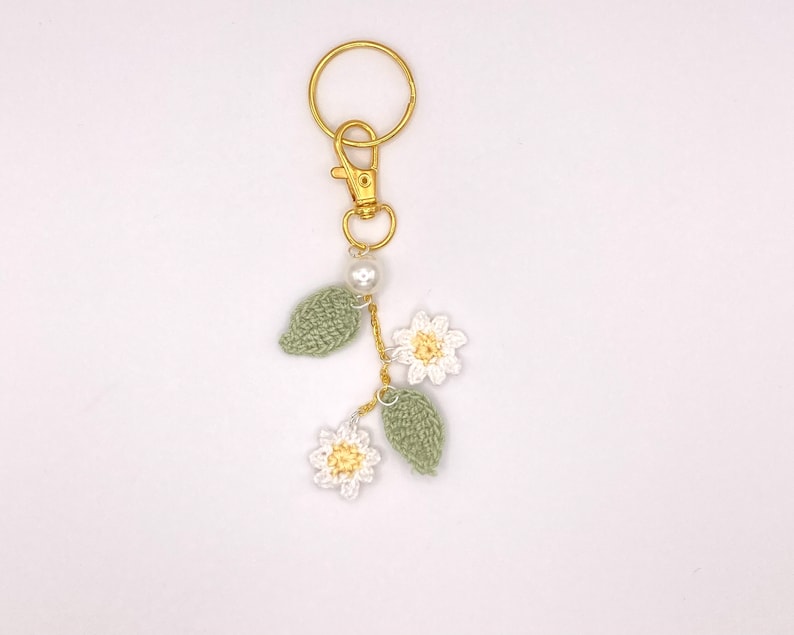 May include: A gold key chain with a crocheted flower and leaf charm. The charm is white and green with a pearl bead.