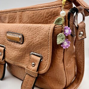 May include: A brown leather shoulder bag with a gold chain and a crocheted flower charm. The charm features purple and white flowers with green leaves.