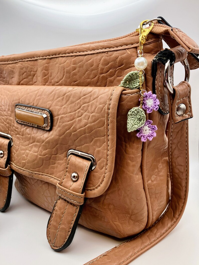 May include: A brown leather shoulder bag with a gold chain and a crocheted flower charm. The charm features purple and white flowers with green leaves.