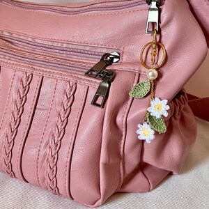 May include: A pink faux leather handbag with a braided design and a crocheted flower keychain with a pearl charm.