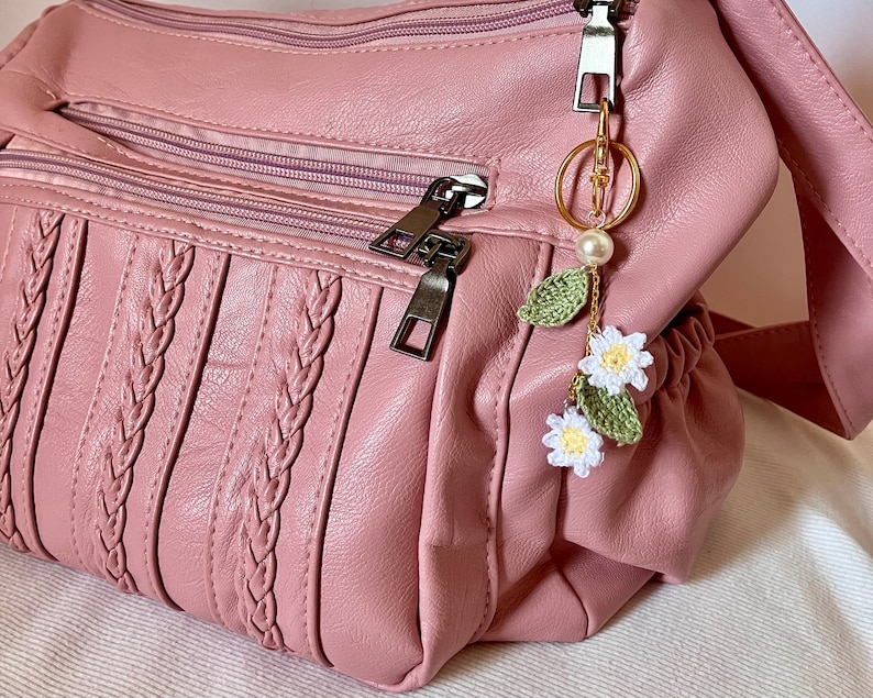May include: A pink faux leather handbag with a braided design and a crocheted flower keychain with a pearl charm.