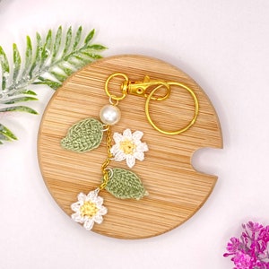 May include: A crocheted flower keychain with a gold key ring and a white pearl bead. The keychain features three white flowers with yellow centers and two green leaves.