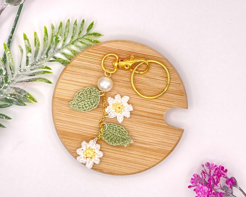 May include: A crocheted flower keychain with a gold key ring and a white pearl bead. The keychain features three white flowers with yellow centers and two green leaves.