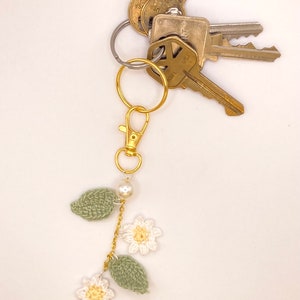 May include: A gold key chain with a crocheted flower and leaf charm. The charm is attached to a gold chain with a pearl bead. The keychain is attached to a set of keys.