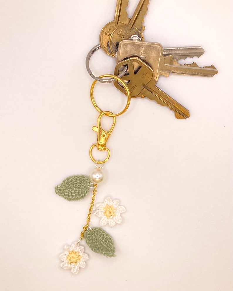 May include: A gold key chain with a crocheted flower and leaf charm. The charm is attached to a gold chain with a pearl bead. The keychain is attached to a set of keys.