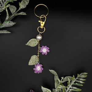May include: A crocheted flower keychain with a gold key ring, a pearl bead, and green leaves. The keychain is hanging against a black background.