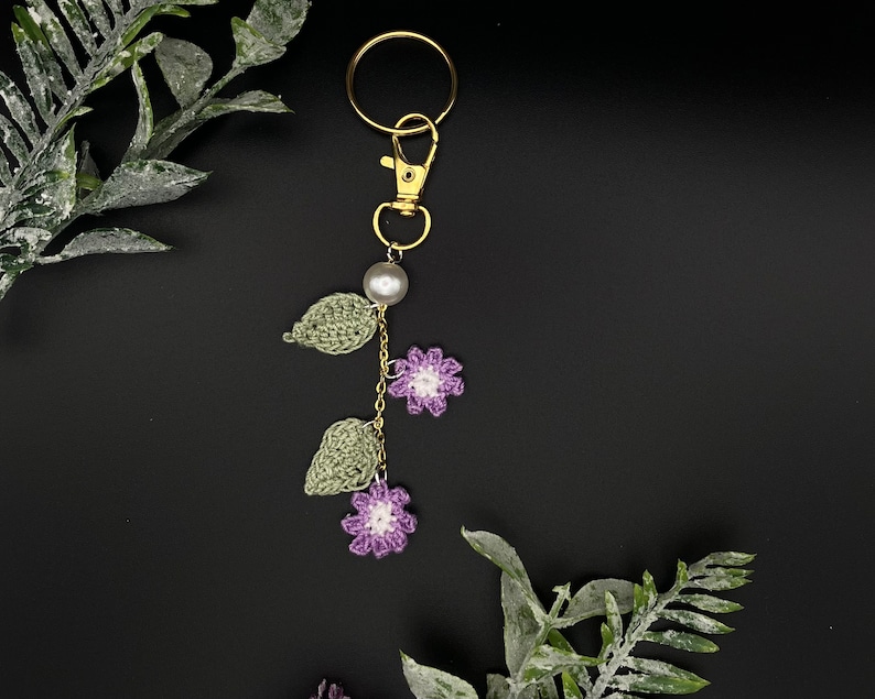 May include: A crocheted flower keychain with a gold key ring, a pearl bead, and green leaves. The keychain is hanging against a black background.