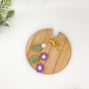 May include: A purple and green crocheted flower keychain with a gold key ring. The keychain is displayed on a wooden surface.