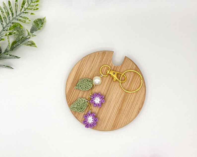 May include: A purple and green crocheted flower keychain with a gold key ring. The keychain is displayed on a wooden surface.